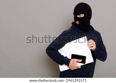 Man steals laptop and phone, runs away and turns back, being of persecution, wears robber mask and black turtleneck, advertising space, isolated over gray background.