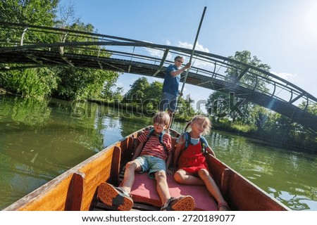 A man is steering a boat down a river under a bridge, with two children seated in the vessel, enjoying a sunny day visiting Oxford, England