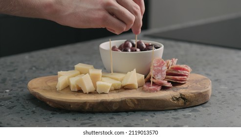 man stick toothpicks in olives, cheese and fuet on olive wood board
