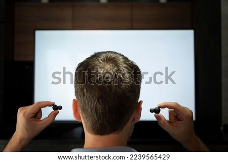 a man sticks headphones in his ear against the background of a TV screen. watching TV with headphones at night.