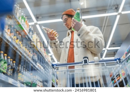 Man in store chooses drink. Guy in supermarket reads label, composition of sodas, chooses, compares prices, looks for benefits. Bottom view from grocery cart. Racks with huge variety of drinking goods