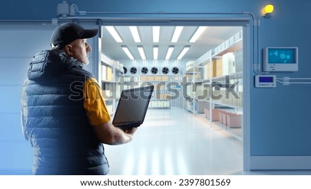 Man storekeeper with laptop. Refrigerated warehouse. Large freezer in factory. Worker near refrigerator door. Industrial refrigerator employee. Man master adjusts refrigeration equipment via computer