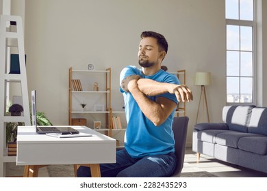 Man stretches during work break. Young man in blue T shirt sitting by desk with laptop computer in home office and doing relaxing exercise to prevent muscle pain, discomfort and stiffness in shoulders