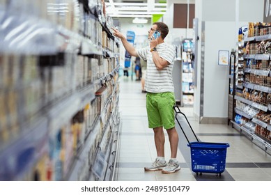 Man in striped shirt and green shorts shops in grocery store aisle while talking on his phone. He pulls blue shopping basket on wheels. - Powered by Shutterstock