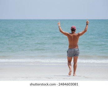 A man in striped shorts and a cap raises both arms in celebration on a sandy beach, expressing joy and victory with the ocean as a backdrop - Powered by Shutterstock