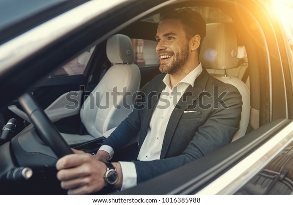 Man of style and status. Handsome young man\
in full suit smiling while driving a\
car