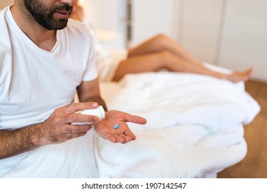 Man Suffering From Erectile Dysfunction Holding Pill In Hand And His Wife In Background. Man Takes Viagra Before Sex, Woman Behind Him In Bed.