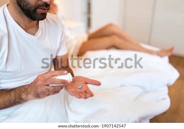 Man Suffering From Erectile Dysfunction Holding\
Pill In Hand And His Wife In Background. man takes viagra before\
sex, woman behind him in\
bed.