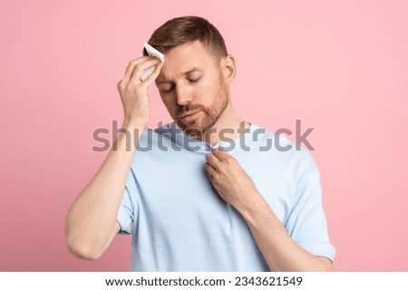 Man suffering from heat stuffiness wiping sweat on forehead with paper napkin isolated on pink background. Exhausted overheated bearded male with closed eyes. Summer hot weather, stuffy room concept. Сток-фото © 