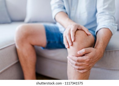 Man suffering from knee pain sitting sofa. A mature man massaging his painful knee. Man suffering from knee pain at home, closeup. Pain knee 