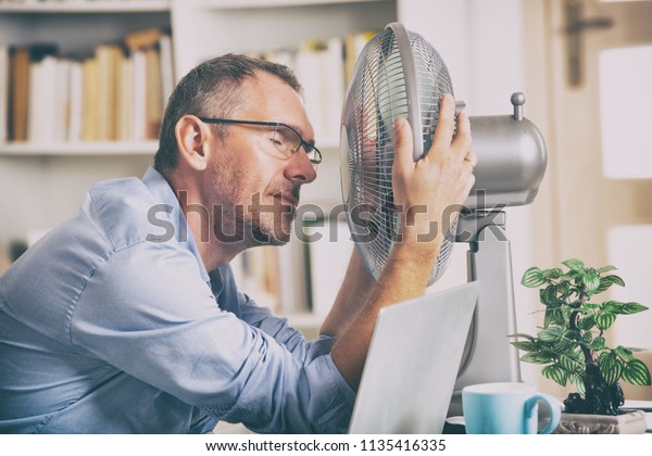 Man suffers from heat while working in the\
office and tries to cool off by the\
fan