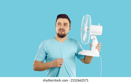 Man suffers from summer heat at home. Guy with broken air conditioner in his house using bad electric fan and sweating. Man in T shirt feeling hot and holding fan with sad face expression, studio shot