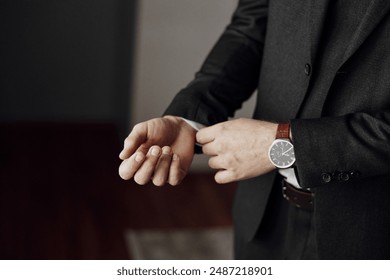 A man in a suit is adjusting his watch. The man is wearing a black suit and a brown watch band - Powered by Shutterstock