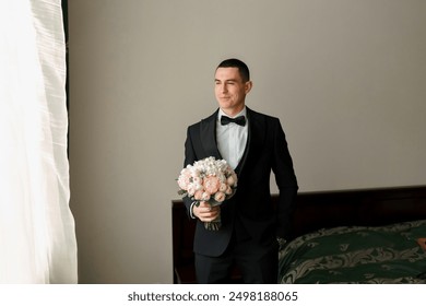 A man in a suit and bow tie stands in front of a bed with a bouquet of flowers in his hand - Powered by Shutterstock