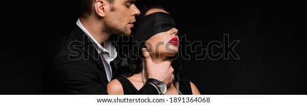 man in suit choking blindfolded woman\
with silk ribbon on eyes isolated on black,\
banner