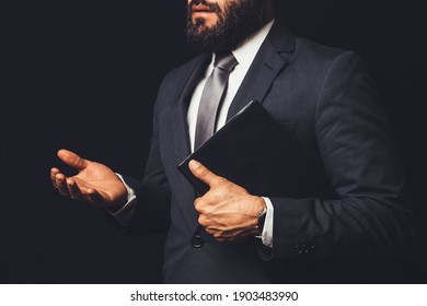 Man In Suit Holding A Bible In His Arm Speaking To Another Person On A Black Background