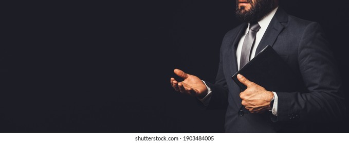 Man In Suit Holding A Bible In His Arm Speaking To Another Person On A Black Background