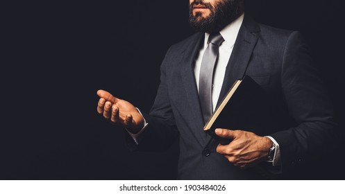 Man In Suit Holding A Bible In His Arm Speaking To Another Person On A Black Background