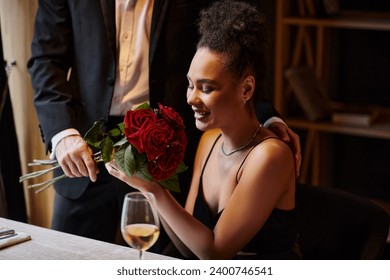 man in suit holding roses near happy young african american woman in restaurant on Valentines Day