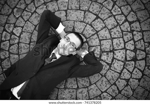 A man in a suit lies on a brick floor\
outdoor. A businessman in a businesslike gray suit in a city\
street. Business man posing on camera\
outdoors\
\
