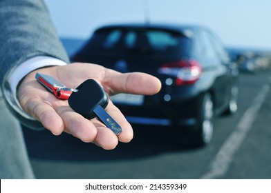 man in suit offering a car key to the observer, with a car in the background