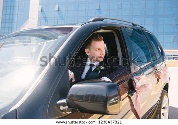 A man\
in a suit sits behind the wheel of a black\
car.