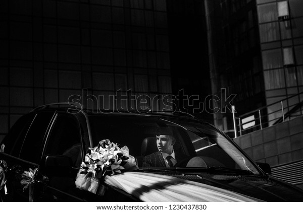 A man\
in a suit sits behind the wheel of a black\
car.