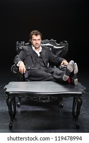 Man In A Suit Sitting In A Chair With His Feet On The Desk, Isolated On A Black Background.