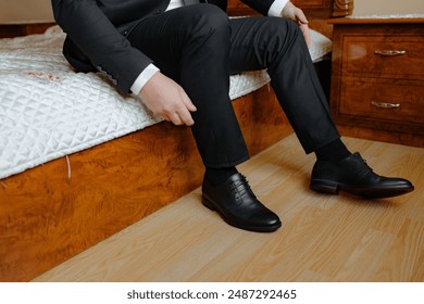 A man in a suit is sitting on a bed with his feet up. He is wearing black dress shoes and is adjusting them. Concept of relaxation and comfort, as the man is taking a break from his formal attire - Powered by Shutterstock