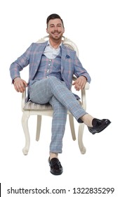 A Man In A Suit Is Sitting On An Expensive Chair. Front View Of A Guy In Stylish Clothes On A Chair. Isolated On White Background. A Rich, Confident Man Smiling Looking Into The Frame.