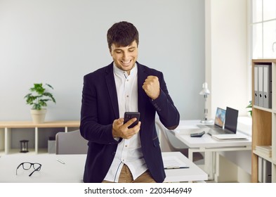 Man In Suit Standing In Company Office, Looking At Mobile Phone, Clenching Fist And Smiling. Successful Business Man Receives Good News At Work. Corporate Worker Happy To Get Money On First Pay Day