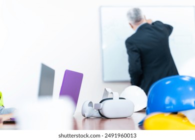 A man in a suit is standing with his back to the camera, writing on a whiteboard. There are two hard hats and a virtual reality headset on the table in front of him.