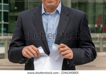 A man in a suit tearing a piece of paper against the background of a modern office building. Concept: termination of contract, invalid document, dismissal from work.