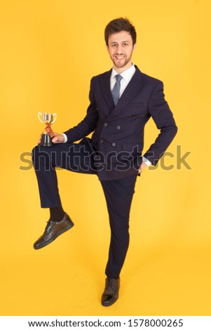 A man in a suit with a tie Handsome looking face with beard In the business man look Holding a trophy on one leg And smiling at you In the yellow background