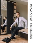 A man in a suit and tie sits on a bed in front of a mirror. He is wearing black dress shoes and a white shirt