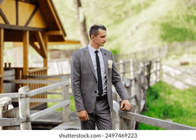 A man in a suit and tie stands on a wooden bridge. He is holding a white flower in his hand - Powered by Shutterstock