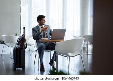 Man With Suitcase Sitting In Airport Waiting Area. Business Man Sitting At Airport Lounge With Laptop Having Coffee And Looking Away.