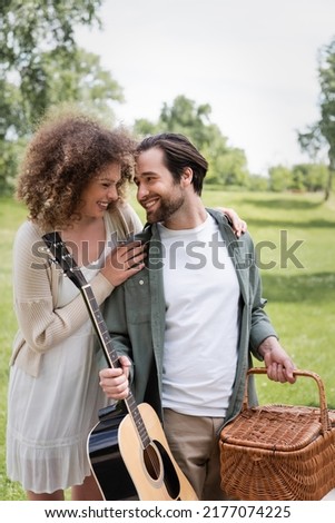 man in summer clothes carrying guitar and wicker basket while looking at happy girlfriend in green park