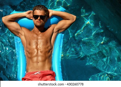 Man Summer Fashion. Beautiful Male With Sexy Body In Swimwear, Fashionable Sunglasses Tanning, Floating In Swimming Pool Water At Relax Spa Resort. Fitness Model With Skin Sun Tan Relaxing On Vacation