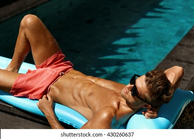 Man Summer Fashion. Beautiful Sexy Male With Fit Body In Swimwear, Fashionable Sunglasses Tanning By Swimming Pool At Resort Relax Hotel. Fitness Model With Healthy Skin Sun Tan Relaxing On Vacation