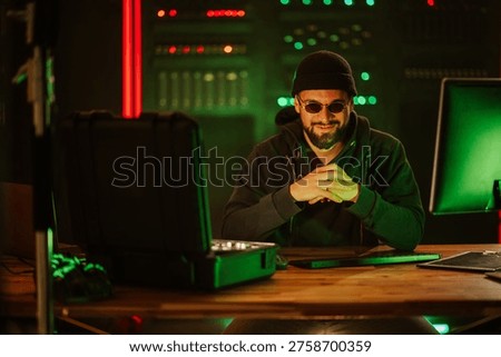 Man in sunglasses and beanie using computer, planning cyberattack in a dark room with green and red lights