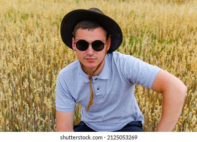 Man In Sunglasses. Portrait Of Farmer Seating In Gold Wheat Field With Blue Sky In Background. Young Man Wearing Sunglasses And Cowboy Hat In Field Examining Wheat Crop. Oats Grain Industry. Closeup.