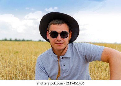 Man In Sunglasses. Portrait Of Farmer Standing In Gold Wheat Field With Blue Sky In Background. Young Man Wearing Sunglasses And Cowboy Hat In Field Examining Wheat Crop. Oats Grain Industry. Closeup.