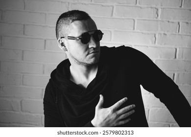 Man in Sunglasses Posing Dramatically Against a White Brick Wall in a Casual Black Outfit.