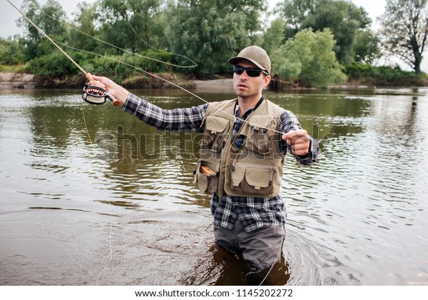 Man in sunglasses
stands in water and fishing. He is preparing fly-fishing. Guy holds
it in one hand and spoon in the other one. Man wears shirt, waders,
vest and cap.