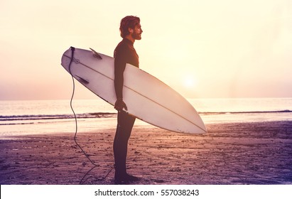 Man surfer carrying his surfboard at sunrise - Hipster male in wetsuit waiting for the high waves on beach - Extreme sport concept - Focus on male silhouette - Matte filter with soft blue vignette - Powered by Shutterstock