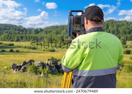 Man surveyor with his back to camera. Specialist in geodesy near forest. Builder surveyor measures plot of land. Man worker with optical theodolite. Surveyor inspects construction site.