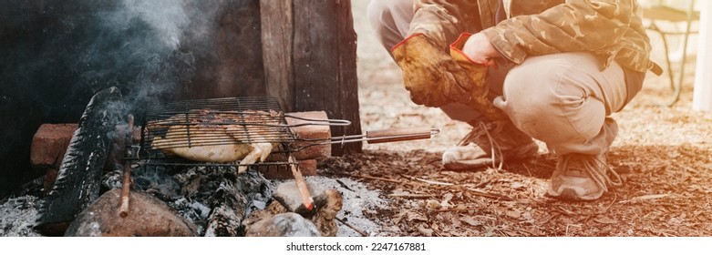 man survivalist cooks roasts chicken meat food are fried on grill on smoldering coals or ember from campfire on ground. barbecue in camping conditions. countryside and wild rustic life. banner. flare - Powered by Shutterstock