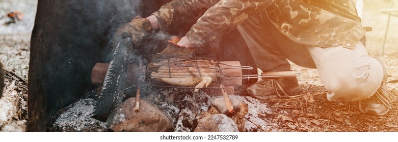 man survivalist cooks roasts chicken meat food are fried on grill on smoldering coals or ember from campfire on ground. barbecue in camping conditions. countryside and wild rustic life. banner. flare - Powered by Shutterstock