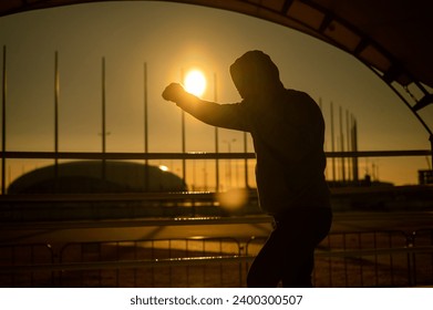 A man in a sweatshirt trains in boxing at the stadium at sunset. Athlete silhouette.